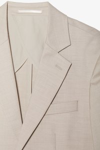 Blazer beige en tissu léger avec une texture lisse. Il présente des revers crantés, une poche poitrine simple et une finition soignée.