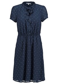 Robe marine en tissu léger, présentant des manches courtes, un col nœud, et des pois blancs. La taille élastique crée une silhouette ajustée.