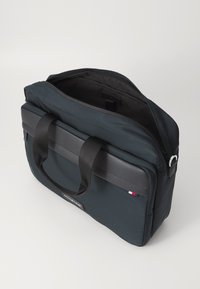 Czarna torba na laptopa z nylonu z płaskim wierzchem, mocnymi uchwytami i szarym akcentem. Posiada zamek błyskawiczny i małą metkę z logo. Wnętrze jest ciemne.