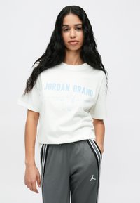 Fiatal nő, aki hosszú fekete hajat visel, fehér Jordan Brand pólót és szürke nadrágot hord, fekete és fehér oldalsó csíkokkal.