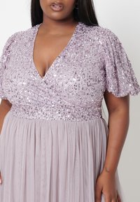 Lila paljettklänning med en djup V-ringning, korta puffärmar och en svävande plisserad kjol. Slät textur och glittrande detaljer.