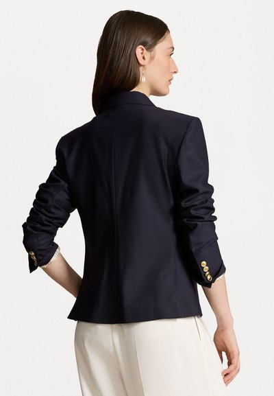 Blazer marine avec un design structuré, featuring des manches retroussées et des accents de boutons dorés sur les poignets. Tissu lisse avec une coupe ajustée.