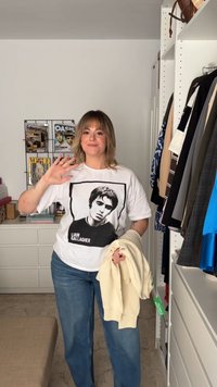 Žena mává jednou rukou, má na sobě tričko s grafikou Liam Gallagher, drží krémový svetr a stojí v místnosti s oblečením a časopisy.