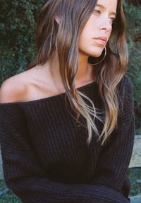 Svart ribbad stickad tröja med avskalat axelkoncept. Långt, vågigt brunt hår, bär stora ringörhängen. Mjuk textur och avslappnad passform.