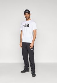 Άσπρο ανδρικό T-shirt με μαύρο λογότυπο, μαύρο παντελόνι με λογότυπο στον μηρό και μαύρα αθλητικά παπούτσια. Απλός σχεδιασμός, άνετη εφαρμογή, ανθεκτικό υλικό.