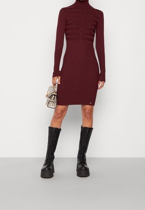 Abito lavorato a maglia bordo bordeaux con texture a coste, colletto alto e maniche lunghe; abbinato a stivali neri con tacco alto al ginocchio e lacci, e una piccola borsa a tracolla con motivo a serpente.