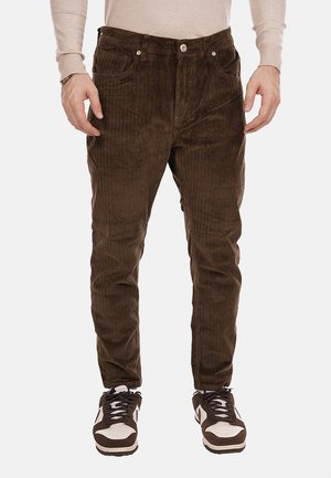 Pantaloni in corduroy marrone con una texture a righe, dotati di cinque tasche, chiusura con bottone e vestibilità affusolata, abbinati a sneaker marroni e bianche.