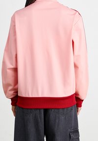 Růžová bunda s hladkým povrchem, kontrastním červeným žebrovaným lemem a rukávy. Disponuje jednoduchým designem bez viditelných log nebo vzorů.