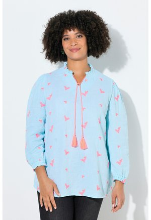 Kobieta o kręconych włosach, ubrana w jasnoniebieską bluzkę z różowymi frędzlami i motywami motyli, stoi na białym tle.
