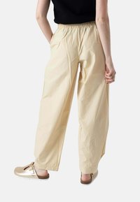 Persona con pantalones sueltos beige con cintura elástica y zapatos dorados sin cordones, de pie frente a un fondo claro y liso.
