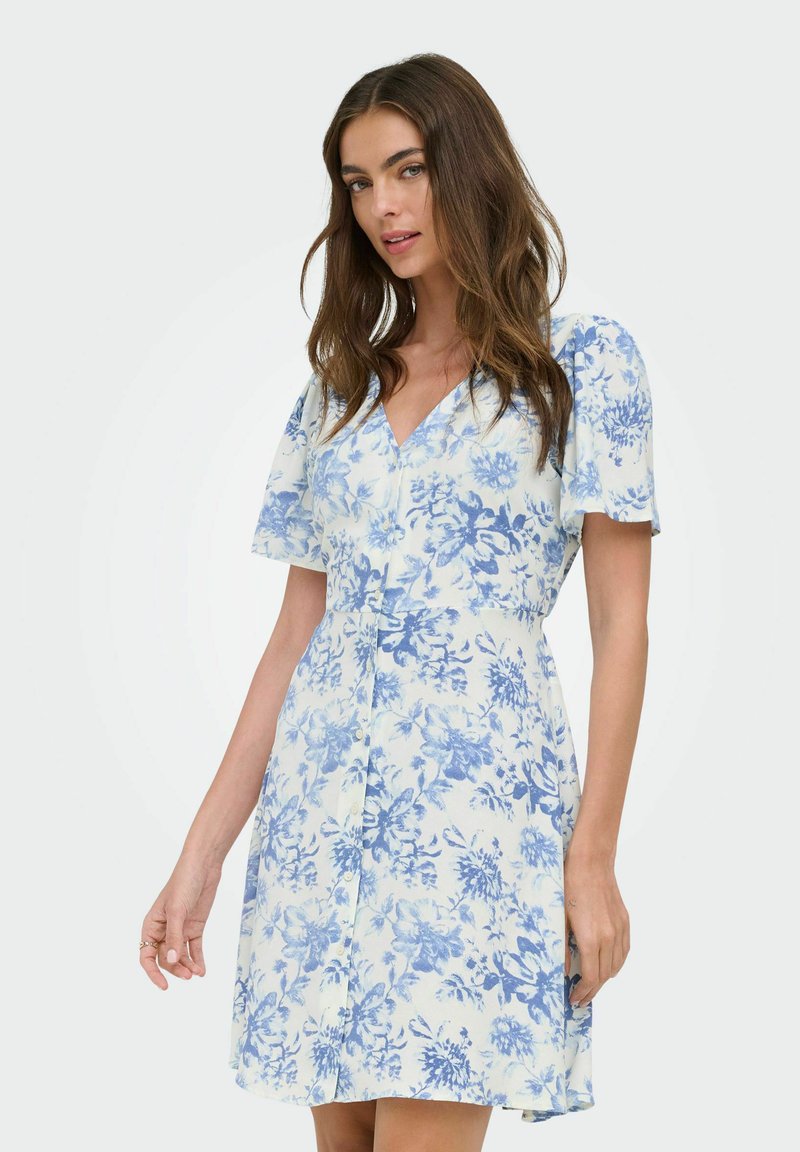Femme portant une robe blanche à manches courtes avec un imprimé floral bleu, debout avec une main légèrement levée, sur un fond uni.