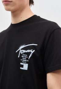 Νέος άνθρωπος που φοράει μαύρο μπλουζάκι Tommy Jeans με ονόματα πόλεων και λογότυπο τυπωμένα στο στήθος, στραμμένος ελαφρώς προς τα αριστερά.