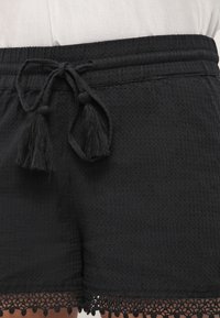 Czarne szorty wykonane z tkaniny o fakturze, z ściągaczem w pasie zakończonym frędzlami. Brzegi mają ozdobne wykończenie z koronką.