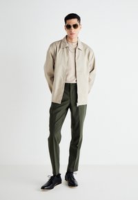 Giacca beige su una camicia crema, abbinata a pantaloni verdi. Scarpe nere, occhiali da sole ovali e un dettaglio in tessuto strutturato sulla giacca.