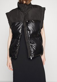 Kobieta w bezrękawniku w kolorze czarnym z pikowanymi panelami na przodzie, nosząca długą czarną suknię, stoi na tle jednolitego jasnego tła.