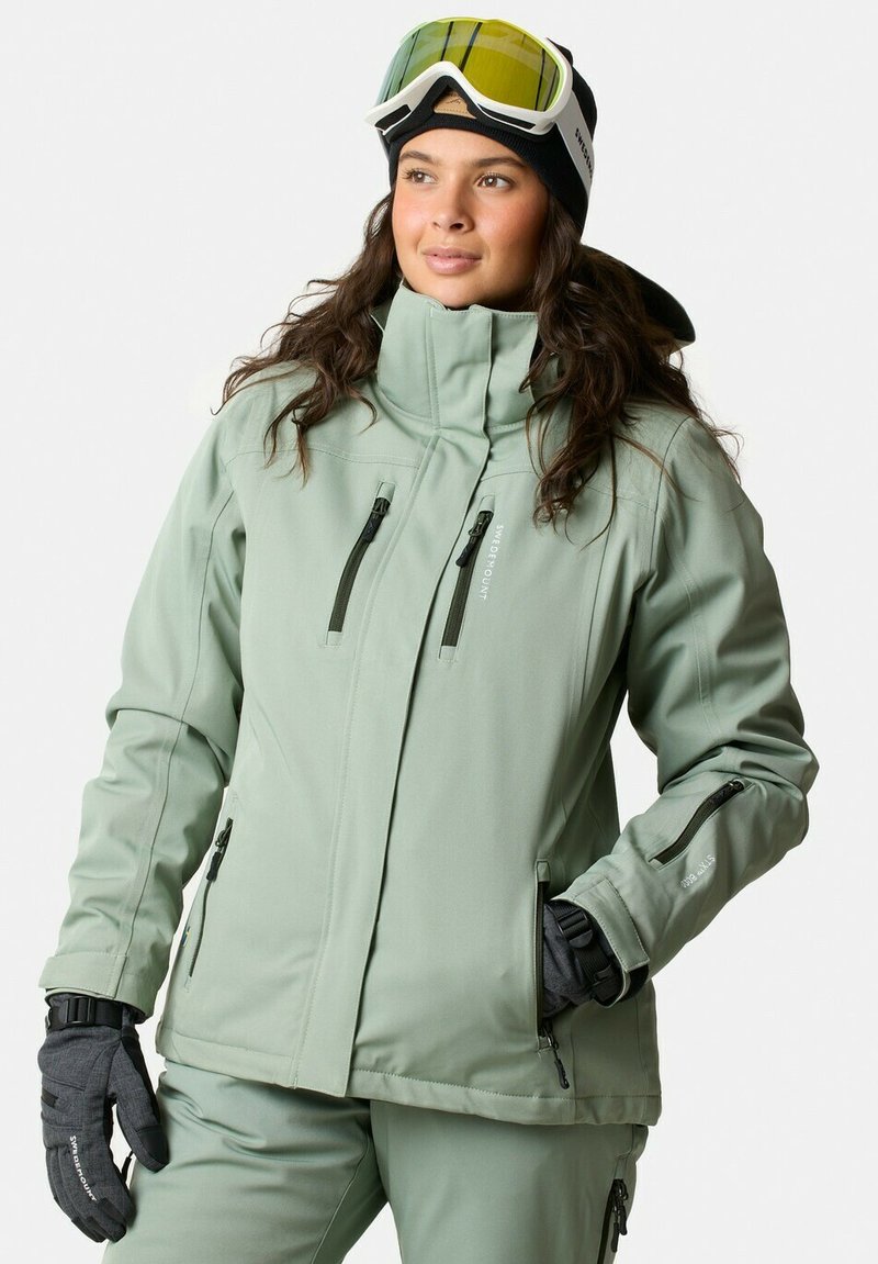 Light green softshell jacket with a high collar, multiple zip pockets, and a zippered cuff. Paired with black gloves and goggles.