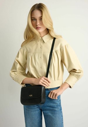 Młoda kobieta o długich blond włosach, ubrana w beżową bluzkę zapinaną na guziki i niebieskie dżinsy, trzymająca czarną torebkę na pasku przewieszoną przez ramię.