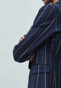 Marynarka marynarska z pionowymi białymi paskami, dopasowany fason, jedna kieszeń na piersi i usztywnione ramiona. Gładka tekstura materiału.