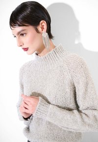 Maglione grigio chiaro con trama testurizzata, collo alto a coste, con fili scintillanti. La modella indossa orecchini lunghi con perline; mani intrecciate in vita.