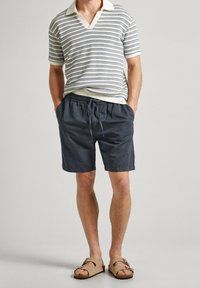 Mężczyzna ubrany w kratkowaną krótko rękawową koszulkę polo, granatowe szorty z paskiem ściągającym i beżowe sandały na klamrę, stojący z rękami w kieszeniach.