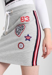 Grauer Rock mit bestickten Aufnähern, darunter ein Herz, ein Stern und Zahlen. Er hat rote und blaue Seitestreifen sowie einen elastischen Bund.