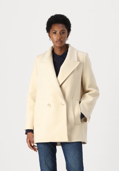 Cremefarvet dobbeltbrystet frakke med store revers, sidelommer og et blødt, tekstureret stof, lagd over en marineblå sweater.