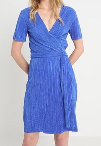 Modrá zavinovací šaty s krátkým rukávem, s vertikálními sklady, hlubokým výstřihem do V a opásáním, které se zavazuje v pase pro přiléhavý siluetu.