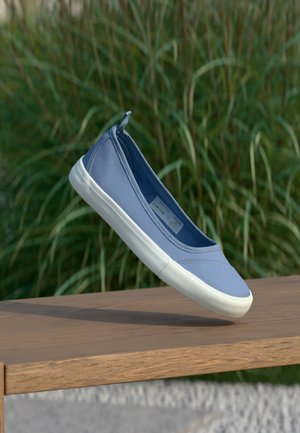 Blauer Slip-On-Leinenschuh mit weißer Gummisohle, schwebend über einer Holzbalkenbank, im Hintergrund grünes Gras.