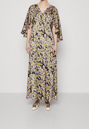 Mujer con un vestido largo de flores en tonos morado, verde y blanco, con mangas amplias y vaporosas, y zapatos blancos puntiagudos con detalles dorados.