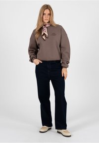 Frau trägt braunen Pullover, dunkle Jeans mit weitem Bein, beige Loafers und einen floralen Seidenschal um den Hals gebunden, steht mit einer Hand in der Tasche.