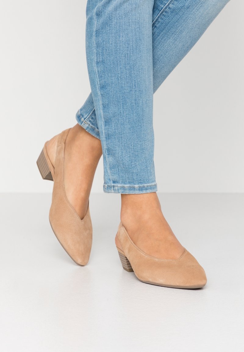 Pieds chaussés de chaussures en daim marron clair à talons bas, bouts pointus et dos ouverts, associés à un jean en denim bleu clair.