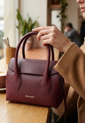Main tenant un sac à main en cuir bordeaux portant l'étiquette "cherry paris" sur une table en bois dans un cadre intérieur chaleureux.