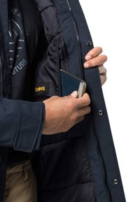 Veste navy avec une fermeture éclair à l'avant, dotée d'une poche intérieure où un smartphone est en train d'être placé. La doublure est lisse et isolante.