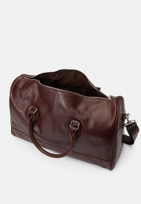 Καφέ δερμάτινη τσάντα duffel με φερμουάρ, διπλές κορυφαίες λαβές, αποσπώμενος ιμάντας ώμου και λεία υφή. Ο εσωτερικός χώρος είναι επενδυμένος με σκούρο ύφασμα.
