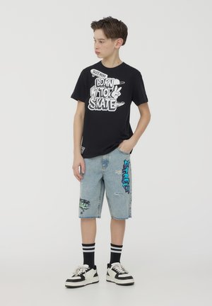 T-shirt nera in cotone con testo grafico bianco "Born to Skate". Pantaloni shorts in denim azzurro chiaro, distrutti, con stampe colorate "Street" e "Cool". Sneakers in bianco e nero.
