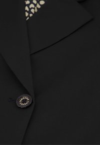 Zwarte blazer met een gladde textuur, voorzien van een gelapte kraag en een ronde zwarte knop met "Fabienne Chapot" in gouden letters, en een gemusterde voering.