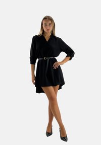 Robe noire à manches longues, décolleté en V, taille cintrée avec une ceinture noire, et un ourlet légèrement allongé à l'arrière. Texture de tissu lisse.