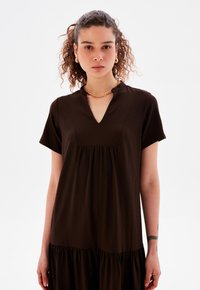 Braunes, kurzärmliges Kleid mit V-Ausschnitt und gerafftem Taillenbereich. Aus strukturiertem Stoff mit ausladendem Saum gefertigt. Goldene Kettenhalskette als Akzent.