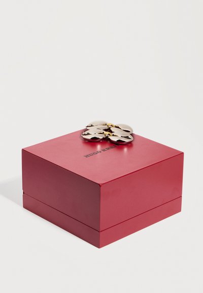 Κόκκινο τετράγωνο κουτί κοσμημάτων με κούμπωμα σε σχήμα χρυσού λουλουδιού και το κείμενο "Hugo K" στην κορυφή, τοποθετημένο σε απλό λευκό φόντο.