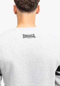 Felpa grigia con collo a giro e strisce nere sulle maniche. Presenta un logo nero "Lonsdale London" sulla parte superiore della schiena.