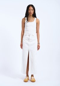 Gonna maxi in denim bianco con spacco frontale e chiusura a bottoni, abbinata a una canotta bianca e sneakers gialle con lacci bianchi.