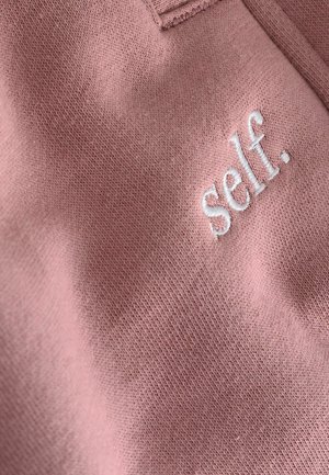 Ružičasta pamučna tkanina s mekanom teksturom koja na prednjoj strani sadrži bijeli izvezeni natpis "self.". Jednostavan i minimalistički dizajn.