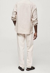 Homme portant une chemise beige à manches longues retroussées, un pantalon beige assorti et des sandales noires, debout de dos face à la caméra.