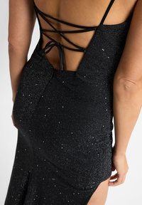 Zwart avondjurk met een glinsterende textuur, open rug en gekruiste schouderbanden. De split aan de zijkant benadrukt de getailleerde silhouet.