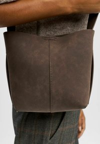 Sac en cuir marron avec une couture centrale, tenu par une personne portant un pantalon à carreaux gris, sur fond uni.