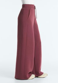 Femeie purtând pantaloni largi, cu talie înaltă, de culoare marsala, cu buzunare laterale și adidași albi, stând pe un fundal simplu, deschis la culoare.