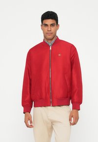 Punane pommijaak, millel on sile tekstuur, soonikkoes käised ja äär. Ees on lukuga ja mõlemal küljel on kaks küljetaskut. Rinnal väike logo.