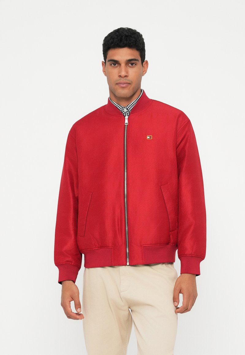 Punane pommijaak, millel on sile tekstuur, soonikkoes käised ja äär. Ees on lukuga ja mõlemal küljel on kaks küljetaskut. Rinnal väike logo.