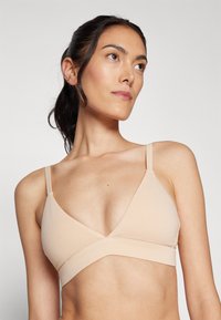 Bralette beige, senza cuciture, con scollatura a V, spalline regolabili e una fascia larga sotto il seno. Texture liscia e morbida senza hardware o decorazioni visibili.