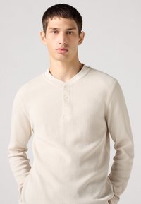 Langermet, beige henley-skjorte med teksturert, ribbet stoff. Har en rund krage og tre knapper i placket ved halsen.